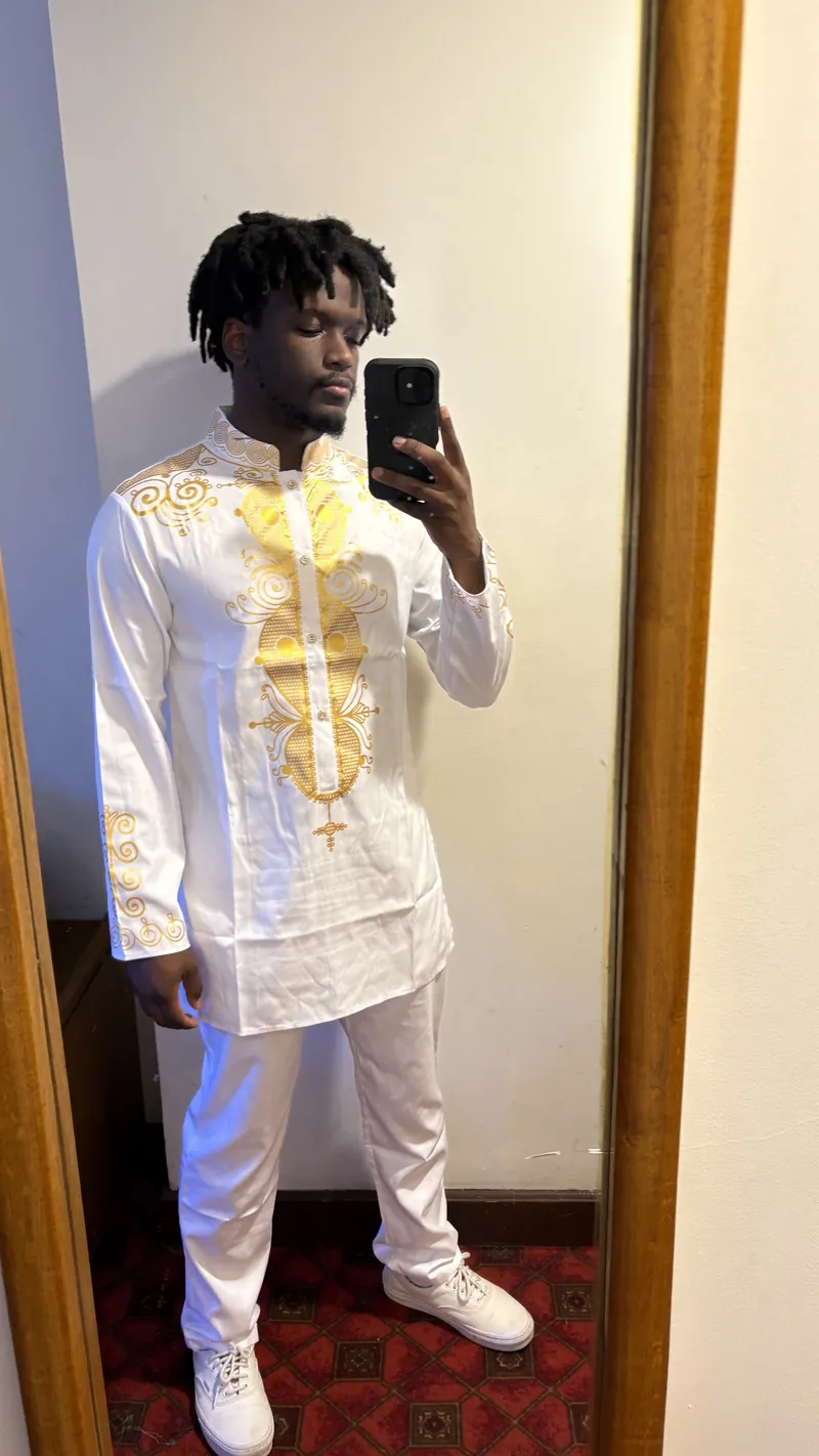 The site owner in a white shirt and trousers with gold embroidery, taking a mirror selfie indoors.