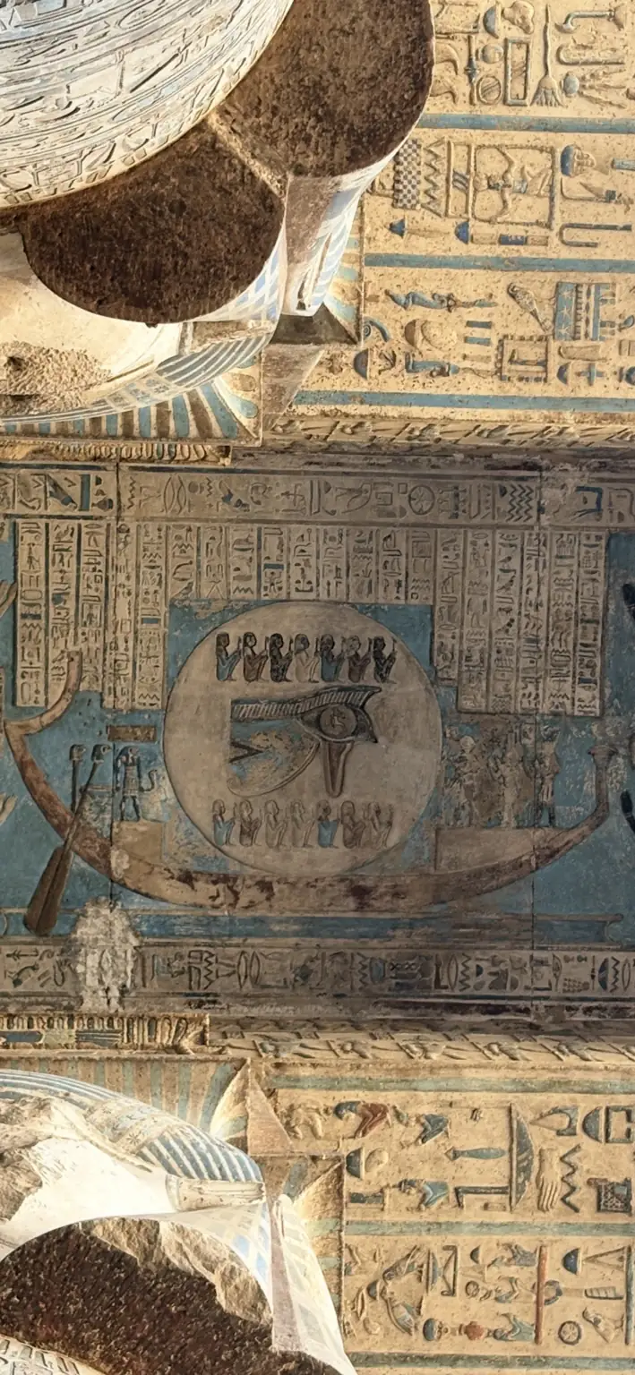 A wide close-up of a painted ceiling field: a large circular Eye sign inside a long boat-shaped composition on a blue ground, with rows of small figures and bands of hieroglyphs above and below, and broken plaster and stone at the edges of the frame.