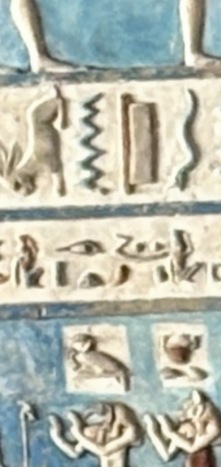 A close-up of a painted relief wall: a row of small hieroglyphs including an eye sign beside a compact group of two rounded shapes above a curved base, with a zigzag sign and a blue-painted field above and below.