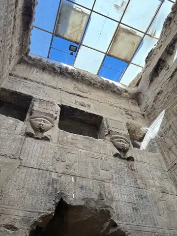 Looking up inside a stone chamber at two carved Hathor faces below a glass skylight.