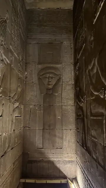 A carved relief of a Hathor face with a frame above it, on the wall of a stone chamber at Dendera, lit in dim light.