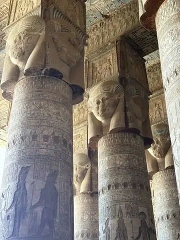 Columns topped with Hathor faces in the hypostyle hall at Dendera, their shafts covered in carved reliefs.